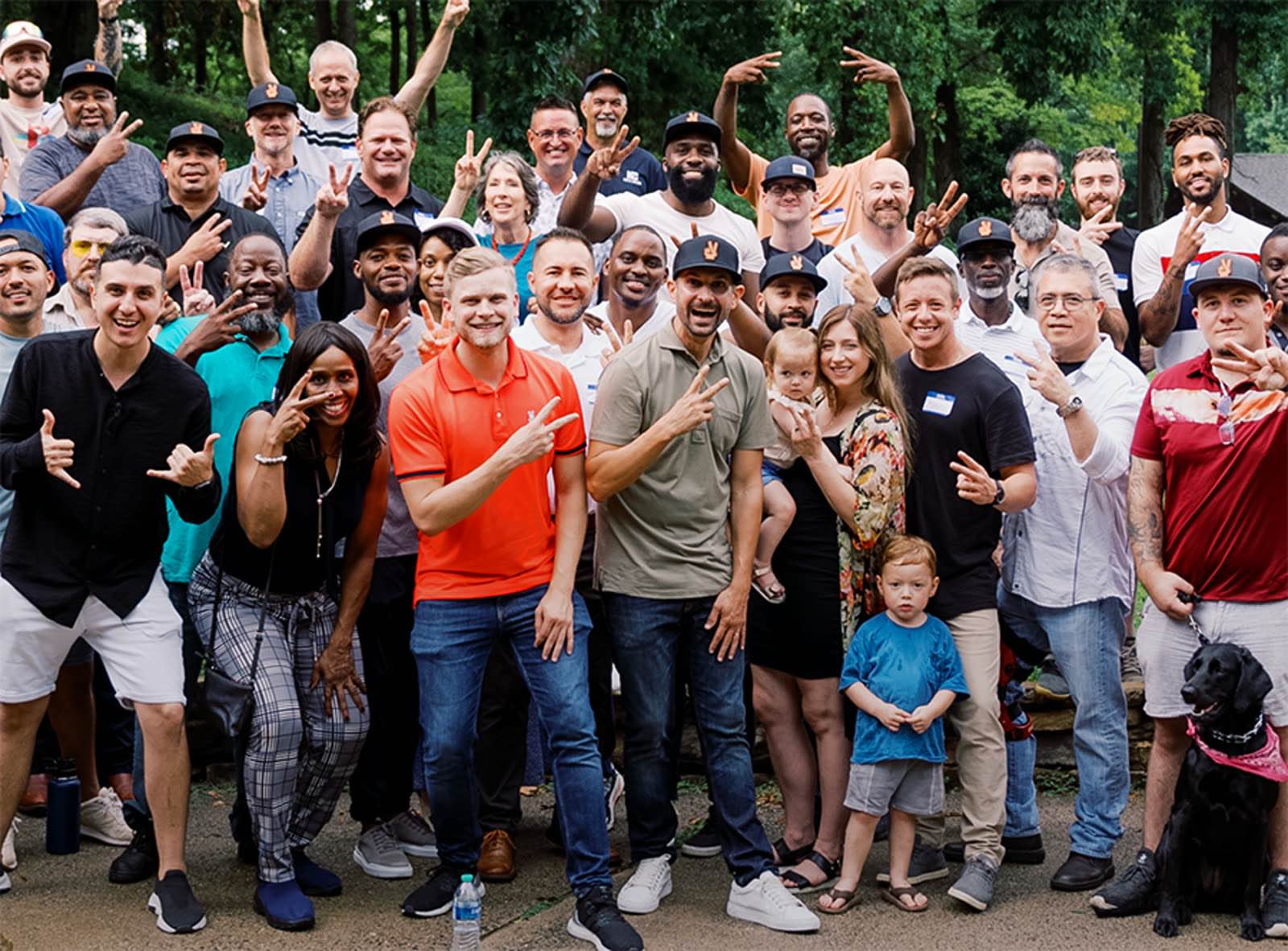 Around forty members and their families stand together outdoors at a SubTo meetup, most holding up the community's two-finger sign.