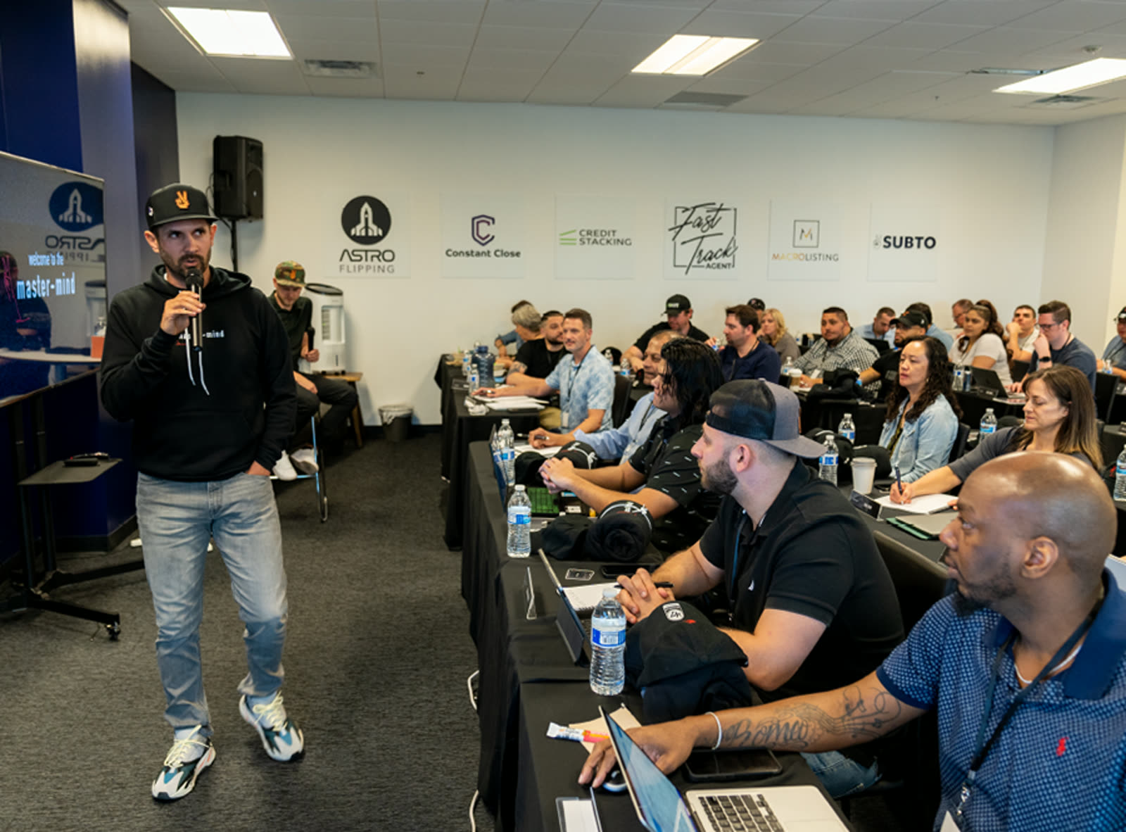 A coach speaks into a handheld mic at the front of a training room while members take notes and work on laptops at long tables.